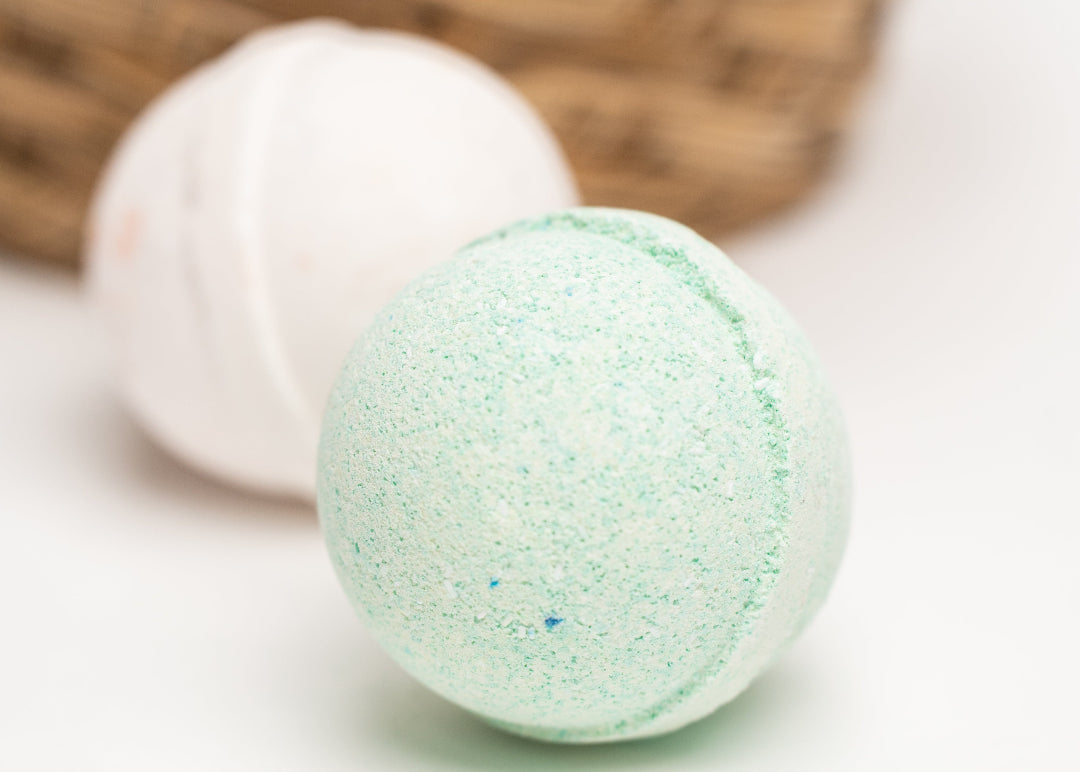Two bath bombs, one green and one white, on a light surface with a wicker basket in the background.